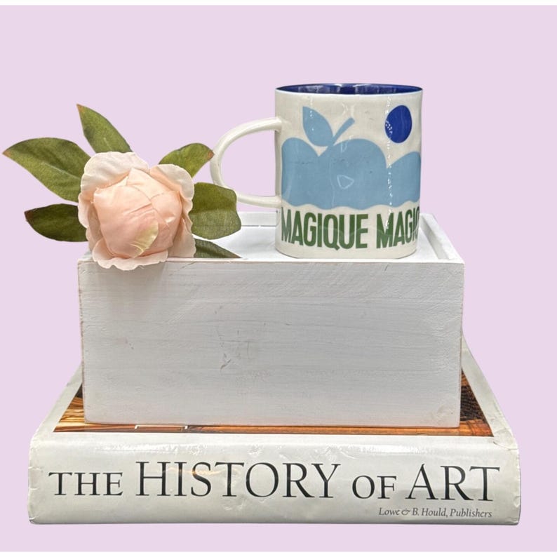 May include: A white ceramic mug with a blue apple design and the words "MAGIQUE MAG" in green. The mug sits on a white tray, next to a pink rose and green leaves. The tray rests on a book titled "THE HISTORY OF ART".