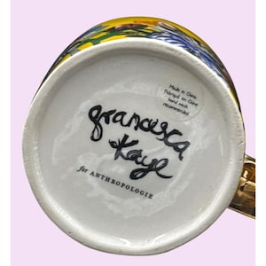 Puede incluir: Una taza de cer&aacute;mica blanca con asa dorada, con un dise&ntilde;o floral colorido. La parte inferior de la taza muestra la firma manuscrita "francesca Kaye" y el texto "for ANTHROPOLOGIE". Una etiqueta dice "Made in China, Fabriqu&eacute; en Chine, hand wash recommended."