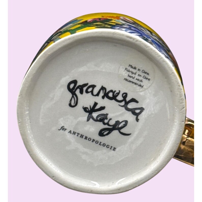 Puede incluir: Una taza de cer&aacute;mica blanca con asa dorada, con un dise&ntilde;o floral colorido. La parte inferior de la taza muestra la firma manuscrita "francesca Kaye" y el texto "for ANTHROPOLOGIE". Una etiqueta dice "Made in China, Fabriqu&eacute; en Chine, hand wash recommended."