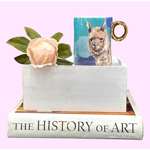 May include: A ceramic mug with a gold handle featuring a painted llama design on a blue background. The mug sits in a white wooden box, next to a peach-colored rose and a book titled "THE HISTORY OF ART".