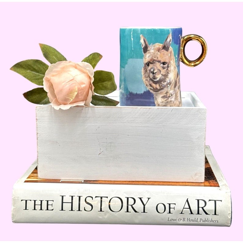 May include: A ceramic mug with a gold handle featuring a painted llama design on a blue background. The mug sits in a white wooden box, next to a peach-colored rose and a book titled "THE HISTORY OF ART".
