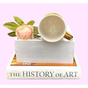 Puede incluir: Una taza de cer&aacute;mica blanca con un asa dorada, apoyada sobre una caja de madera blanca. Una rosa rosa y hojas verdes se colocan junto a la taza. La taza est&aacute; colocada encima de un libro titulado "THE HISTORY OF ART".