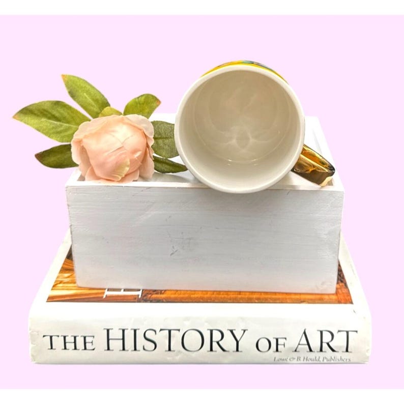 Puede incluir: Una taza de cer&aacute;mica blanca con un asa dorada, apoyada sobre una caja de madera blanca. Una rosa rosa y hojas verdes se colocan junto a la taza. La taza est&aacute; colocada encima de un libro titulado "THE HISTORY OF ART".