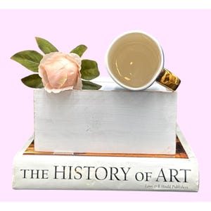May include: A white ceramic mug with a gold handle and a blue rim sits atop a white wooden block, next to a pink rose and green leaves. The block rests on a book titled "THE HISTORY OF ART."