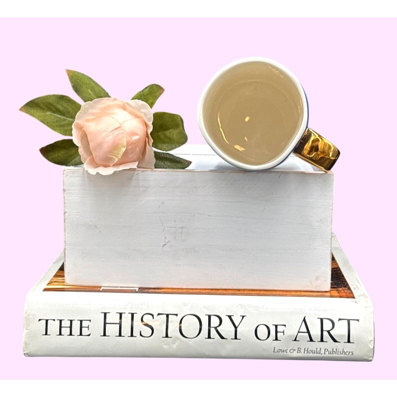 May include: A white ceramic mug with a gold handle and a blue rim sits atop a white wooden block, next to a pink rose and green leaves. The block rests on a book titled "THE HISTORY OF ART."