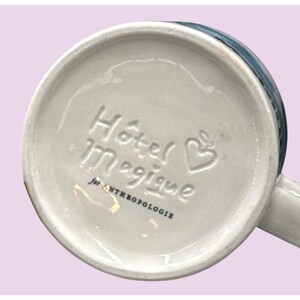 May include: A white ceramic mug with the words "H&ocirc;tel Magique" and a heart-shaped design embossed on the bottom. The text "for ANTHROPOLOGIE" is also present. The mug has a blue rim and a white handle.