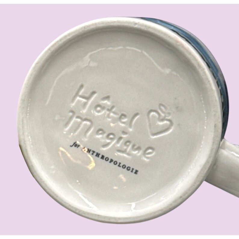May include: A white ceramic mug with the words "H&ocirc;tel Magique" and a heart-shaped design embossed on the bottom. The text "for ANTHROPOLOGIE" is also present. The mug has a blue rim and a white handle.