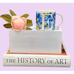 May include: A white ceramic mug with the letter "F" in blue floral print, sits on a white wooden box. A pink flower and green leaves are in the box. The box rests on a book titled "THE HISTORY OF ART".