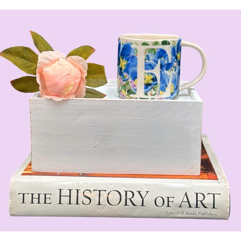 May include: A white ceramic mug with the letter "F" in blue floral print, sits on a white wooden box. A pink flower and green leaves are in the box. The box rests on a book titled "THE HISTORY OF ART".