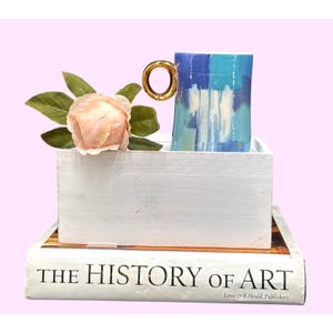May include: A blue and white abstract art mug with a gold handle sits in a white wooden box, next to a pink rose and green leaves. The box rests on a book titled "THE HISTORY OF ART". The mug's design features shades of blue and white.