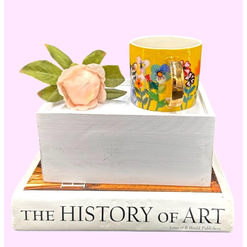 Puede incluir: Una taza de cer&aacute;mica amarilla con un asa dorada, adornada con un dise&ntilde;o floral colorido. La taza est&aacute; sobre una bandeja de madera blanca, junto a una flor color melocot&oacute;n y hojas verdes. La bandeja descansa sobre una pila de libros, incluido uno titulado "THE HISTORY OF ART".