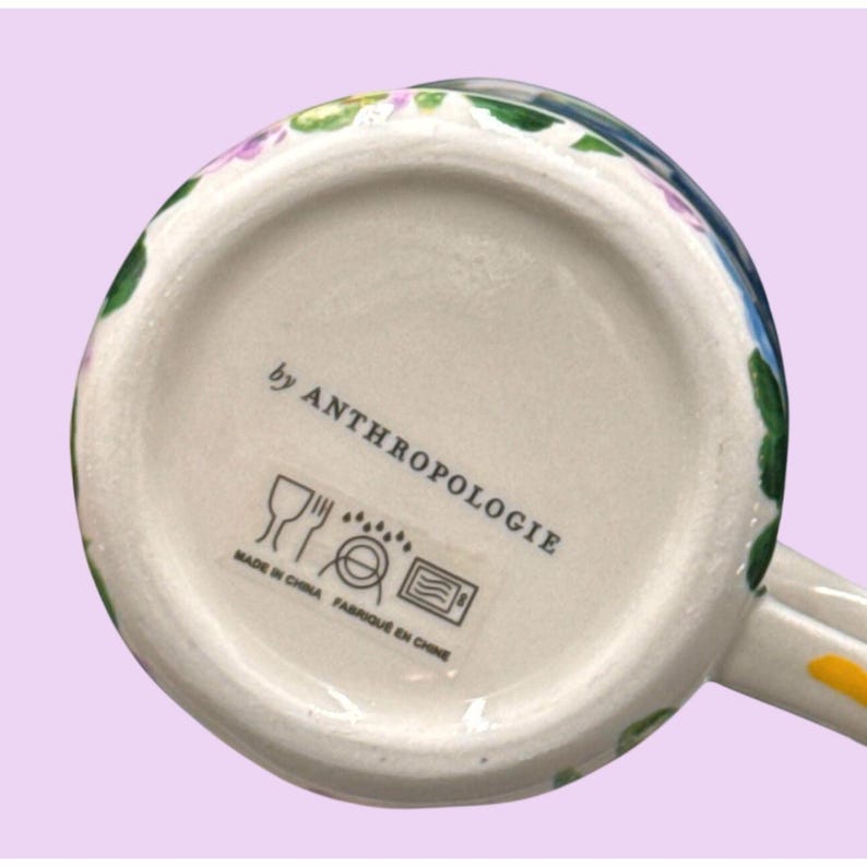 May include: White ceramic mug base with floral design and the text "by ANTHROPOLOGIE". The base of the mug has a stamp that says "MADE IN CHINA FABRIQUE EN CHINE" with symbols for dishwasher and microwave safe.