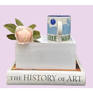 May include: A white mug with blue and green designs and the word "MAGIQUE" is on a white box with a pink flower and a book titled "THE HISTORY OF ART". The mug has a blue interior.