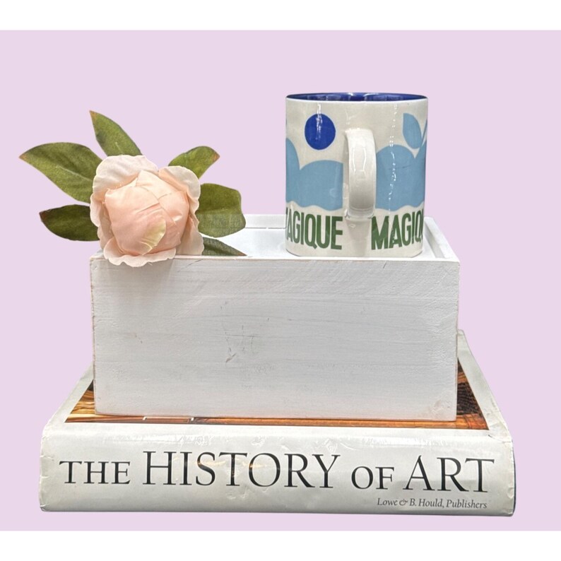 May include: A white mug with blue and green designs and the word "MAGIQUE" is on a white box with a pink flower and a book titled "THE HISTORY OF ART". The mug has a blue interior.