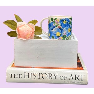 May include: A ceramic mug with a floral pattern of blue flowers, yellow centres, and green leaves. The mug is on a white wooden box, next to a pink flower and a book titled "THE HISTORY OF ART". The background is a pale purple.