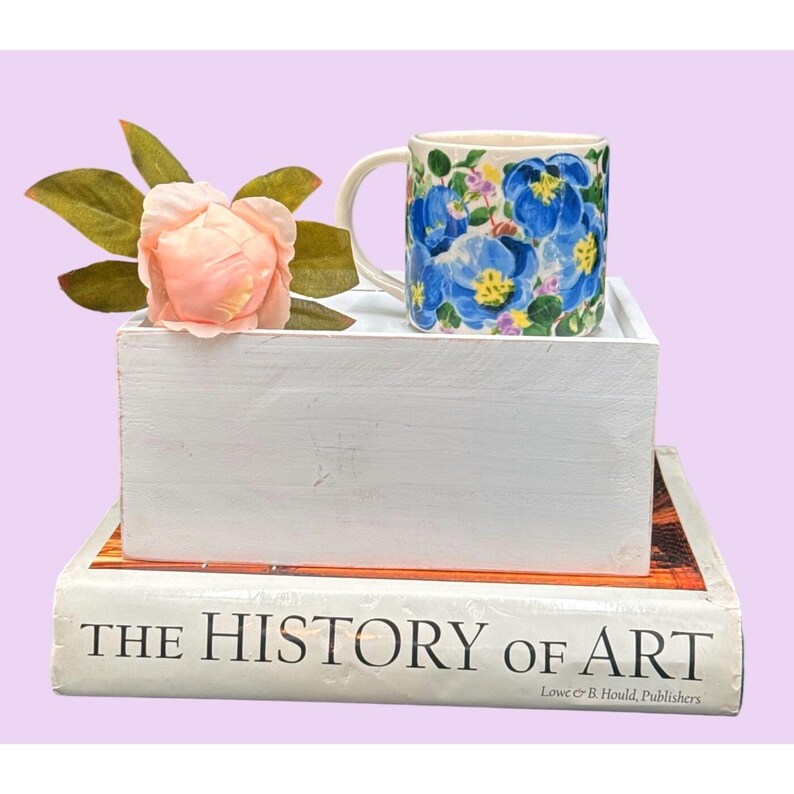 May include: A ceramic mug with a floral pattern of blue flowers, yellow centres, and green leaves. The mug is on a white wooden box, next to a pink flower and a book titled "THE HISTORY OF ART". The background is a pale purple.