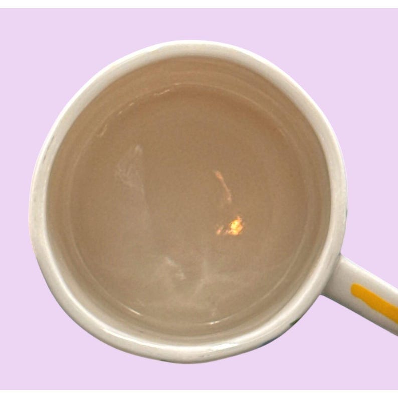 May include: An overhead shot of a white ceramic mug with a light brown interior. The handle is white with a yellow stripe. The mug is set against a light purple background.