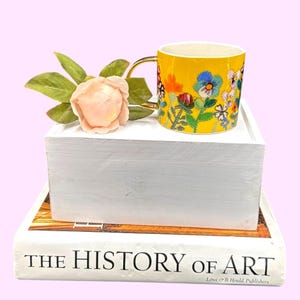 Puede incluir: Una taza de cer&aacute;mica amarilla con asa dorada y dise&ntilde;o floral. La taza est&aacute; decorada con coloridas flores de estilo bordado. La taza est&aacute; sobre un bloque blanco, junto a una rosa rosa y hojas verdes. El libro de abajo dice "THE HISTORY OF ART".