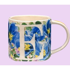 May include: A ceramic mug with a white handle and a floral design. The mug features blue flowers and green leaves, with the white letter "F" prominently displayed. The background is a light purple.