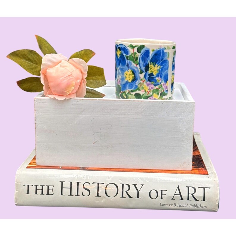 May include: A ceramic mug with blue floral patterns is displayed on a white wooden box, accompanied by a pink flower and green leaves. The box is placed on a book titled "THE HISTORY OF ART". The background is light purple.