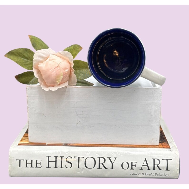 May include: A white ceramic mug with a navy blue interior and rim, resting on a white wooden block. A light pink rose and green leaves are next to the mug. The mug is placed on a book titled "THE HISTORY OF ART".