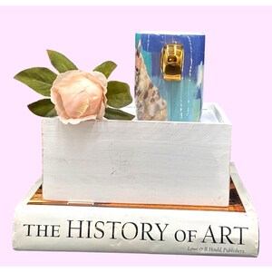 May include: A blue and white ceramic mug with a gold handle, featuring a giraffe design, sits in a white wooden box. A peach-colored rose and a book titled "THE HISTORY OF ART" complete the arrangement.