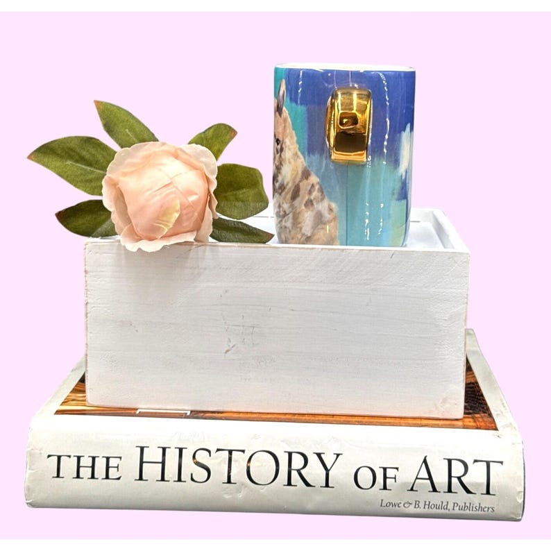 May include: A blue and white ceramic mug with a gold handle, featuring a giraffe design, sits in a white wooden box. A peach-colored rose and a book titled "THE HISTORY OF ART" complete the arrangement.