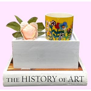 Puede incluir: Una taza de cer&aacute;mica amarilla con un dise&ntilde;o floral, colocada sobre una caja de madera blanca. Una flor rosa artificial y hojas verdes est&aacute;n junto a la taza. La caja est&aacute; sobre un libro titulado "THE HISTORY OF ART".