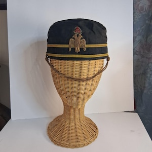 Puede incluir: Una gorra negra con ribete dorado y negro, con un emblema dorado de un águila bicéfala y un sol rojo. La gorra se exhibe en una forma de cabeza de mimbre tejido, mostrando su diseño y detalles.