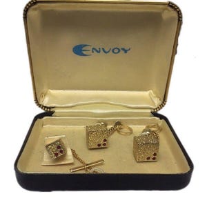 May include: A set of gold-toned dice-shaped cufflinks and a matching tie bar with red accents, presented in a black and cream-colored Envoy branded jewelry box. The dice feature a textured surface with small red dots representing the numbers.