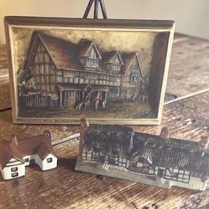 May include: A collection of miniature house figurines and a framed relief sculpture. The relief depicts a detailed Tudor-style building with figures. Two small houses feature brown roofs and white walls. The framed piece is propped on a small easel.