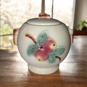 Puede incluir: Azucarero de cerámica blanca con tapa, con un diseño en relieve de manzanas rojas y hojas verdes. El cuenco tiene dos asas pequeñas y un pomo marrón en la tapa. Diseño de estilo vintage.