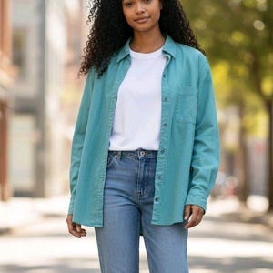 May include: A person wearing a teal button-down shirt over a white t-shirt and blue jeans. The shirt has a single chest pocket. The person is walking on a paved path with buildings and trees in the background.