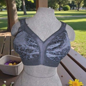 Puede incluir: Un sujetador gris con un estampado floral sobre un maniquí. El sujetador tiene un diseño de cuello en V profundo y tirantes ajustables. El estampado floral es de un tono gris más oscuro, creando un contraste con el fondo gris claro.