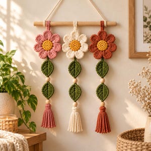 May include: A wall hanging featuring three crocheted flowers in pink, cream, and terracotta, each with a yellow center. Below each flower are two green crocheted leaves, a wooden bead, and a matching tassel, all suspended from a wooden rod.