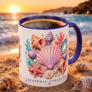 May include: A white ceramic mug with a dark blue interior and handle, featuring a colorful graphic of various seashells, a starfish, and pearls. The text "SEASHELL COLLECTOR" is printed in dark blue at the bottom of the mug's exterior surface.