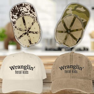 May include: Four baseball caps are displayed. Two caps have a cow print and camouflage pattern. The other two caps are off-white and tan, with the text "Wranglin' feral kids" in black. The caps are on a wooden surface.