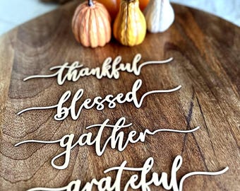 May include: Four white wooden script cutouts featuring the words thankful, blessed, gather, and grateful arranged on a round wooden board with three small decorative pumpkins in orange, yellow, and white colors positioned at the top.