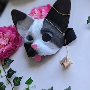 May include: A handmade cat mask with black and white fur, blue eyes, and a pink nose and tongue. A gold chain is attached to the ears, and a small, fluffy toy hangs from the chain. The mask is designed to be worn.