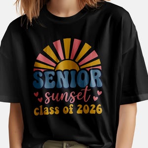 May include: Black t-shirt featuring a colorful graphic design. The design includes the words "SENIOR sunset class of 2026" in a retro font, with a sunset and heart accents. The shirt is made of a soft material.
