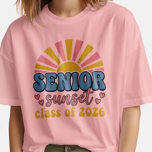 May include: A light pink t-shirt featuring a retro design with the words "SENIOR sunset class of 2026". The design includes a sunburst graphic with pink, yellow, and blue colors. The shirt is a casual style.
