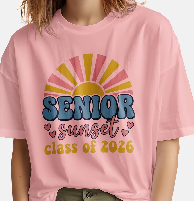 May include: A light pink t-shirt featuring a retro design with the words "SENIOR sunset class of 2026". The design includes a sunburst graphic with pink, yellow, and blue colors. The shirt is a casual style.