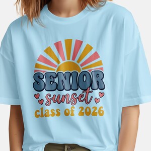 May include: Light blue t-shirt featuring a retro sunset design with the words "SENIOR sunset class of 2026". The design includes a sunburst in yellow and pink, with the text in blue and orange. Small pink hearts accent the word "sunset".