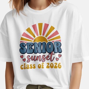 May include: White t-shirt featuring a retro design with the words "SENIOR sunset class of 2026." The design includes a stylized sunset in shades of pink, yellow, and blue, with heart accents. The shirt is a casual, comfortable style.