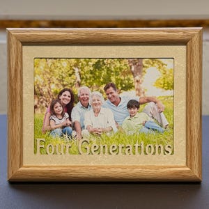May include: A rectangular wooden picture frame with a family photo. The frame has a light brown finish and the text "Four Generations" cut out at the bottom. The photo shows a family of six sitting on the grass.