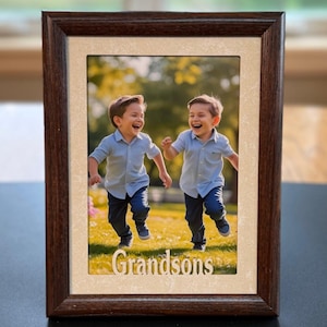 May include: A rectangular, dark brown wooden picture frame with a cream-colored mat. The frame holds a photograph of two young boys running and laughing in a grassy field. The word "Grandsons" is displayed in white letters at the bottom of the frame.