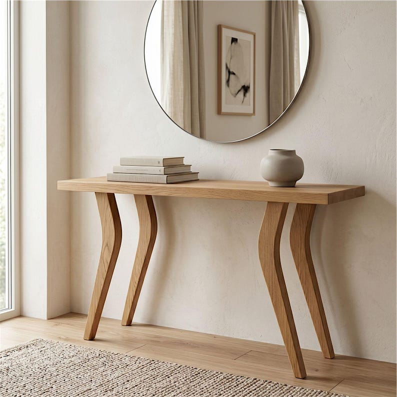 Op de afbeelding: Een licht houten console tafel met een rechthoekig blad en gebogen poten. De tafel is voorzien van een stapel boeken en een kleine grijze vaas. Een ronde spiegel hangt aan de muur boven de tafel.