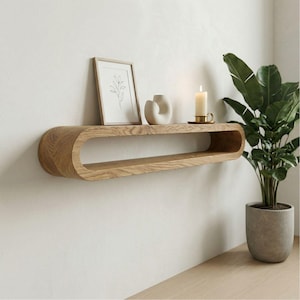 May include: A wall-mounted, oval wooden shelf in a light oak finish. It displays a framed botanical print, a white ceramic vase, and a lit candle. A potted plant is positioned to the right of the shelf.