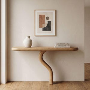 Op de afbeelding: Een moderne, minimalistische interieurscène met een lichte houten console met een gebogen poot. Boven de tafel hangt een ingelijste abstracte kunstprint. Een witte vaas en gestapelde boeken staan op de tafel. Het kunstwerk heeft bruine, zwarte en beige geometrische vormen.