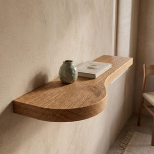Peut inclure: Une étagère flottante en bois avec un bord incurvé est fixée à un mur beige. Un petit vase rond vert et un livre avec une couverture de couleur claire reposent sur l'étagère. L'étagère est en bois clair.